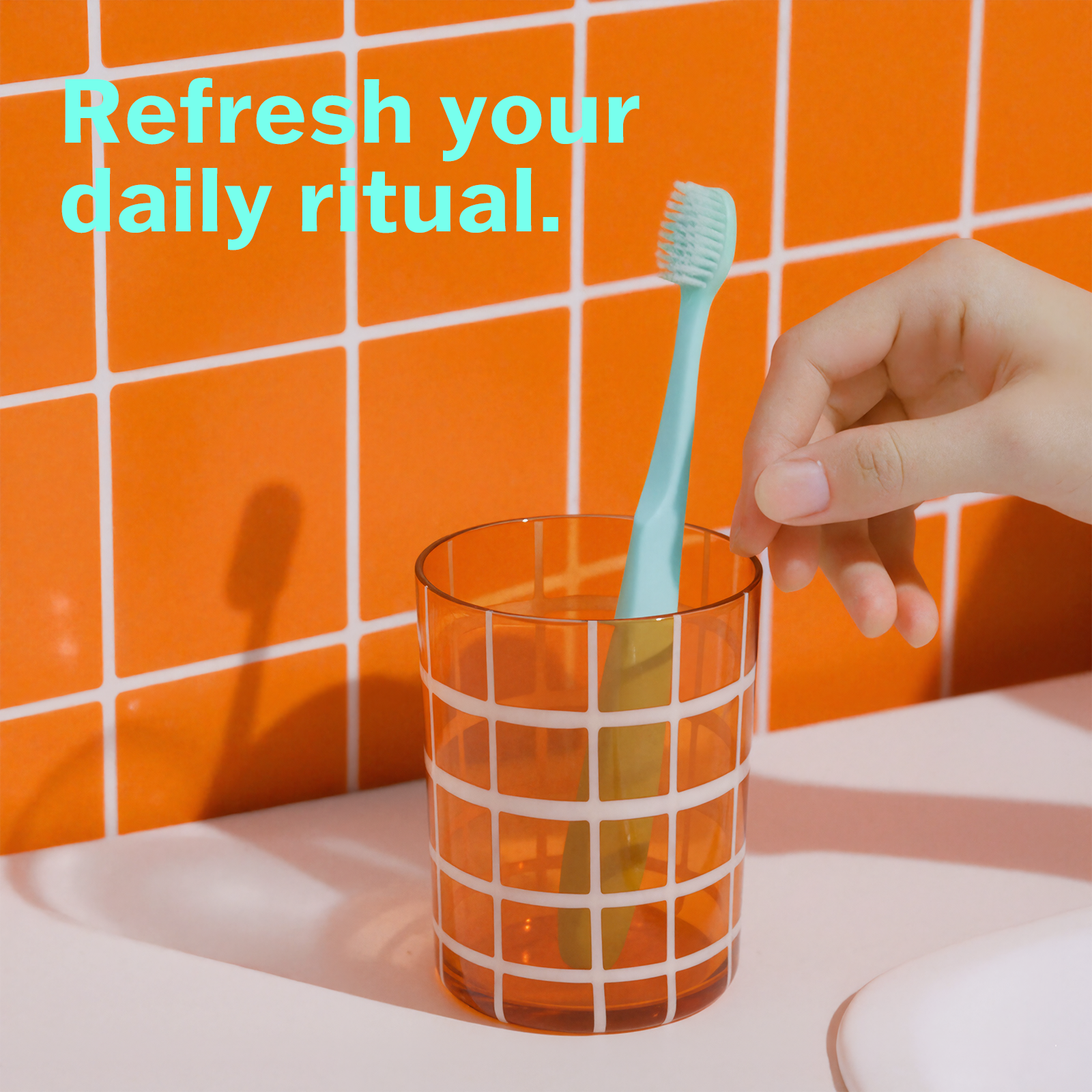 Hand placing a green toothbrush into an orange glass with an orange tiled wall background.