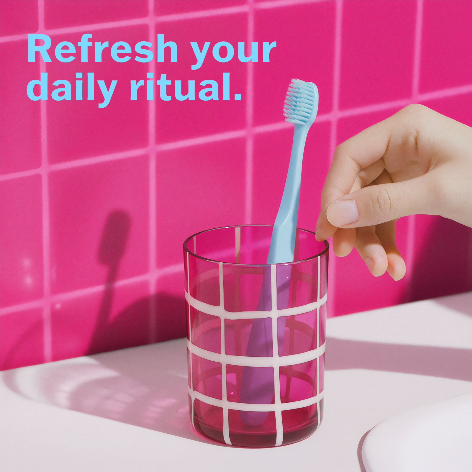 Hand placing a blue toothbrush into a pink glass with a pink tiled wall background.