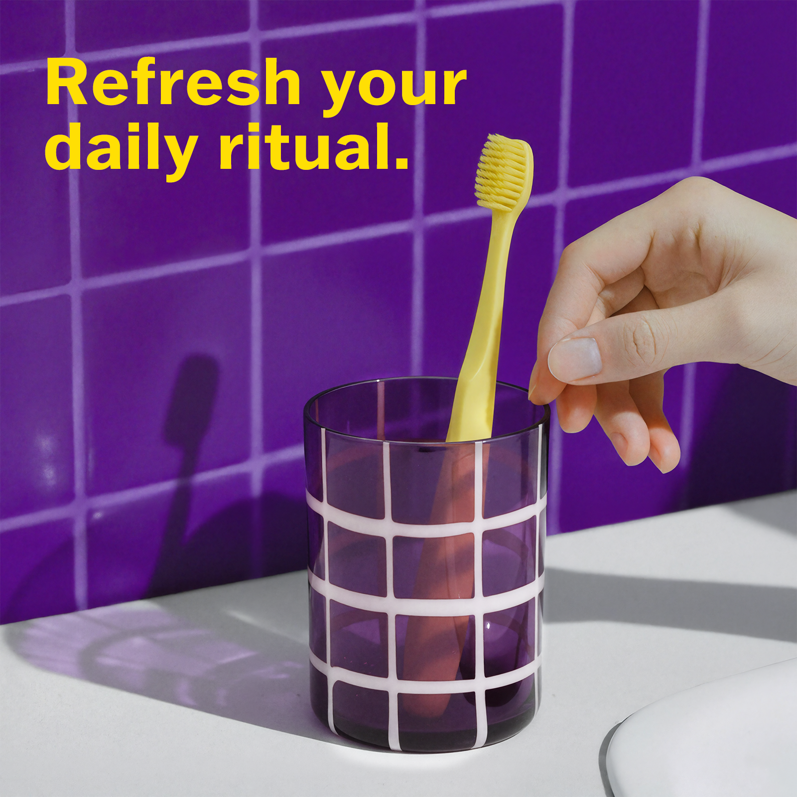 Hand placing a yellow toothbrush into a purple glass with a purple tiled wall background.